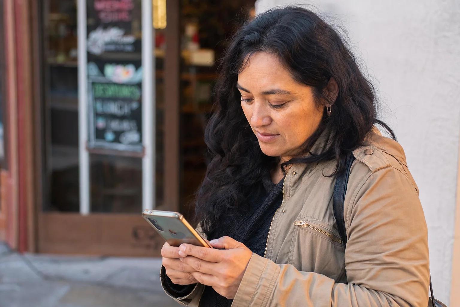 A Latina woman reads a message on her smartphone outside a small business storefront in the late-afternoon light.