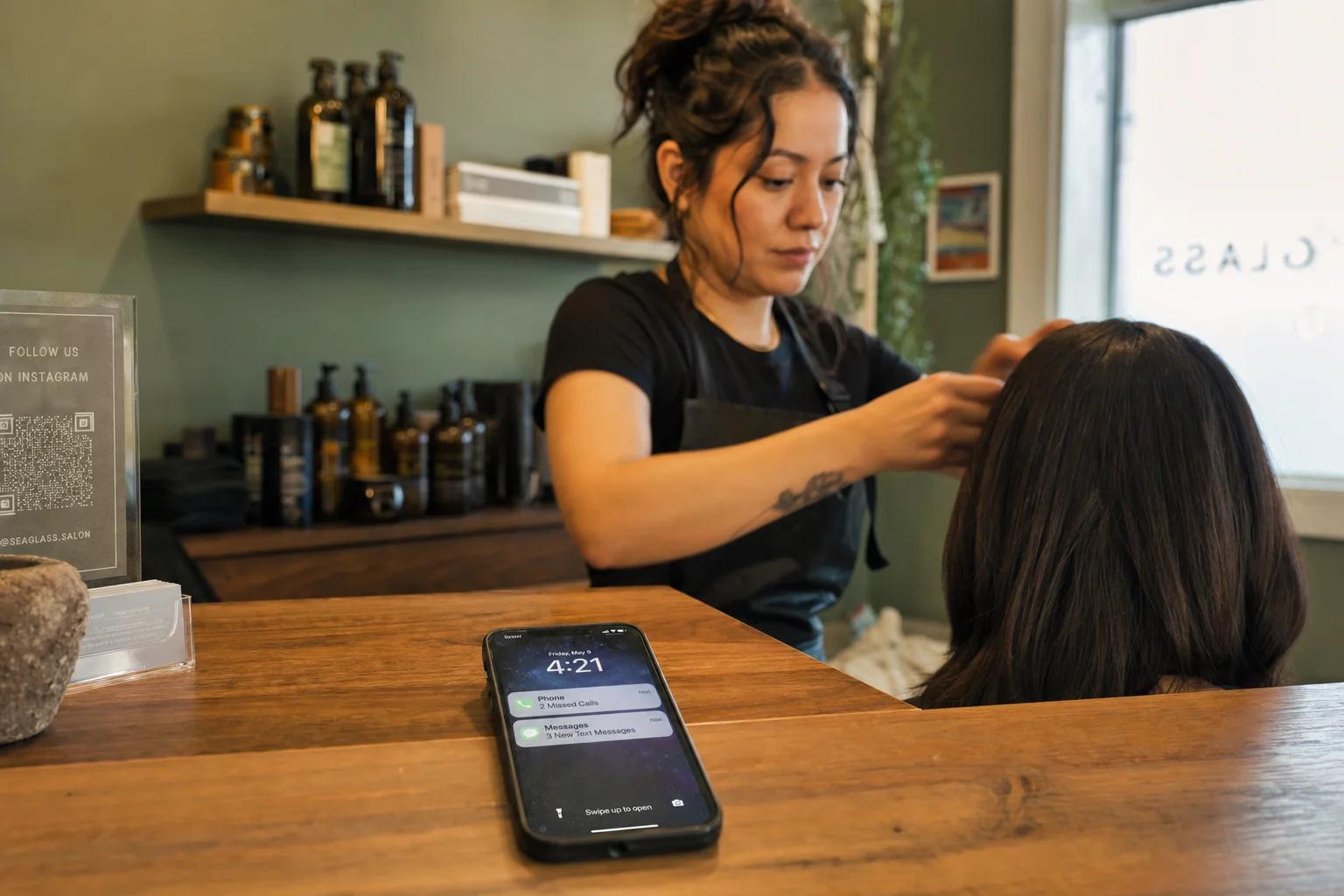 A smartphone on a salon counter shows missed call and SMS notifications while a stylist works on a client's hair in the background.